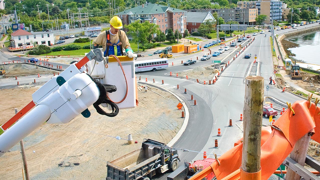 A powerline technician in overalls and a hard hat stands in a utility truck's bucket and approaches a utility pole.