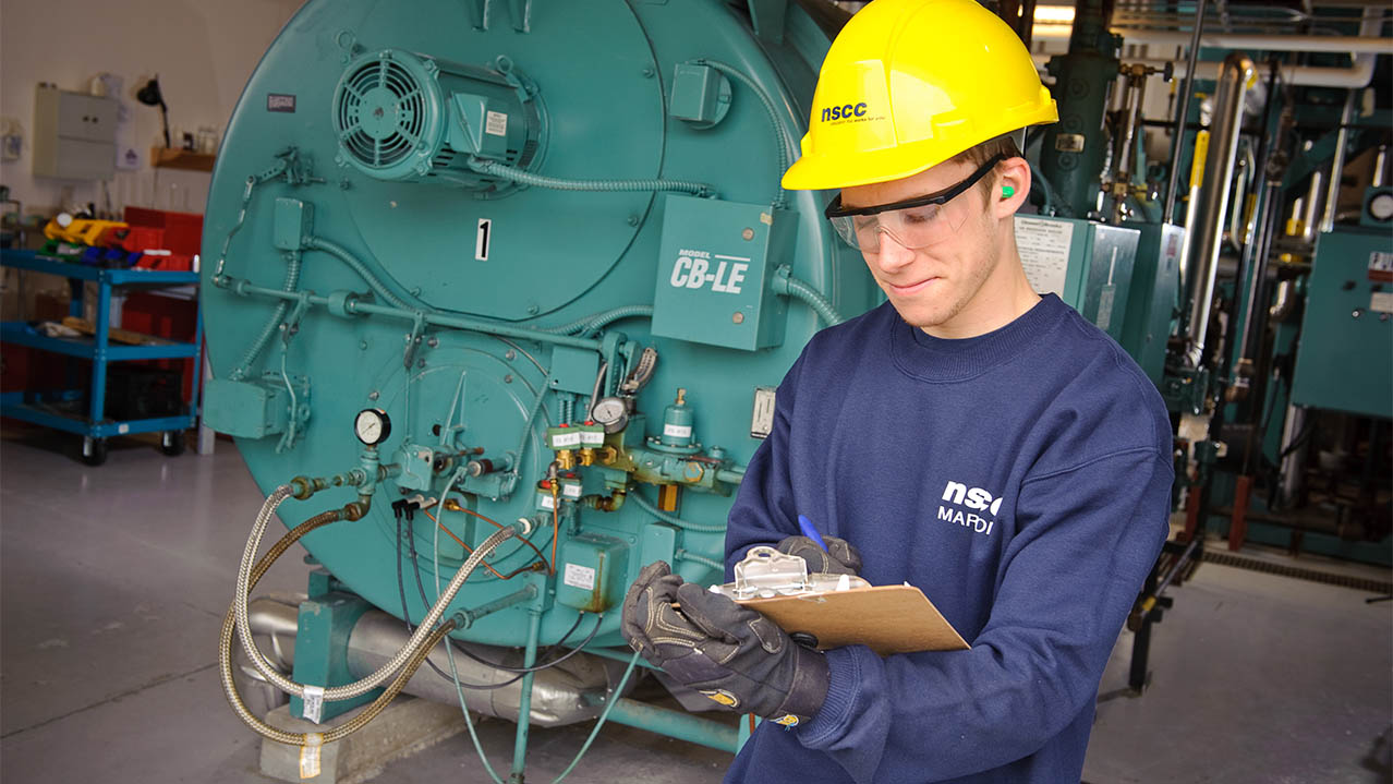 A man in a blue NSCC shirt and yellow NSCC hard hat stands in front of a large piece of equipment and writes on a clipboard.