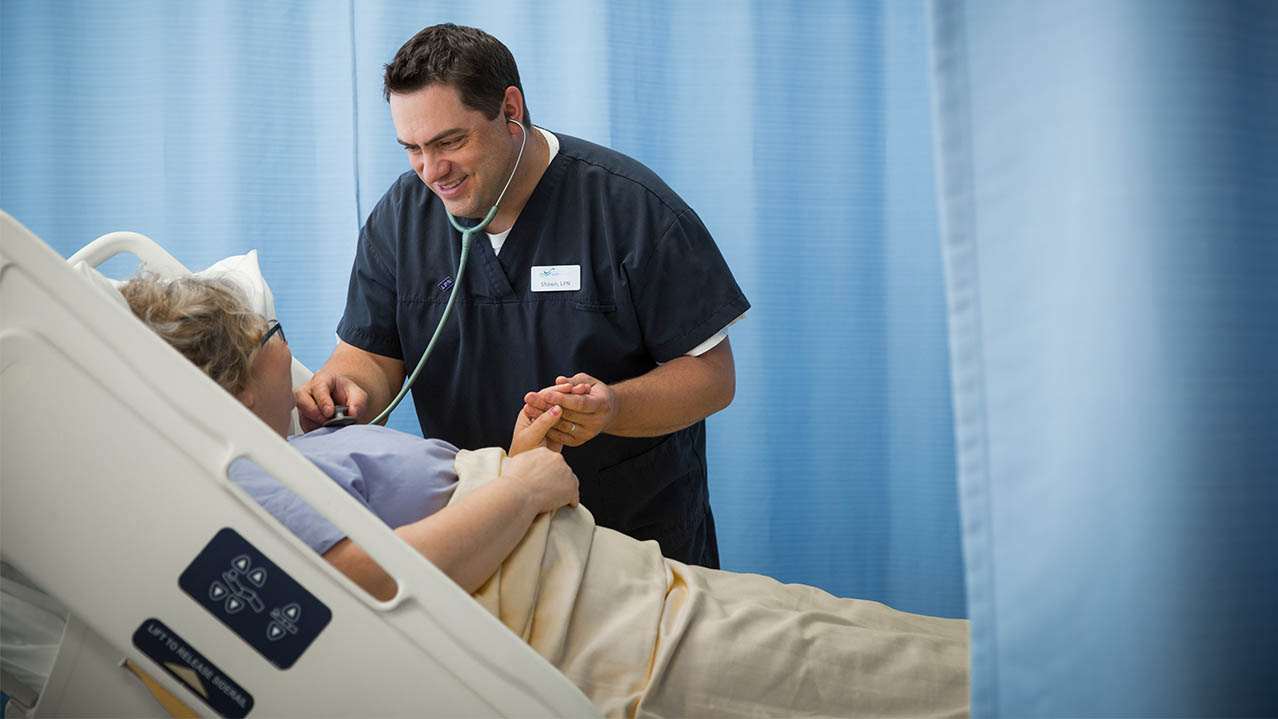 A nurse wearing scrubs smiles while standing near a patient's bed. He holds one of the patient's hands in his and uses a stet