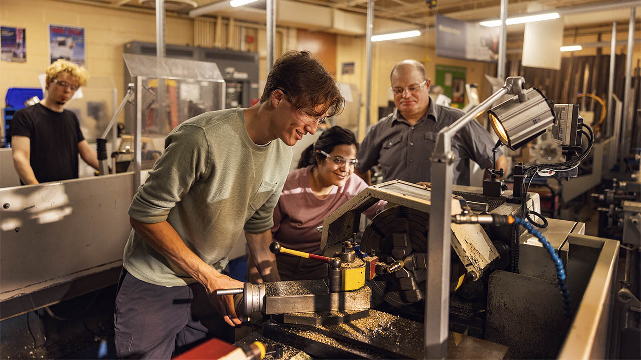 Three students are seen in safety glasses working with their machines in a workshop setting while their teacher watches over 