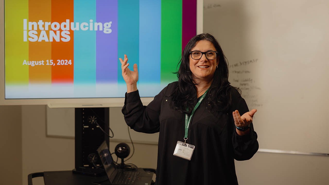 A Public Relations graduate in glasses and a black shirt stands and presents in front of a colourful screen.