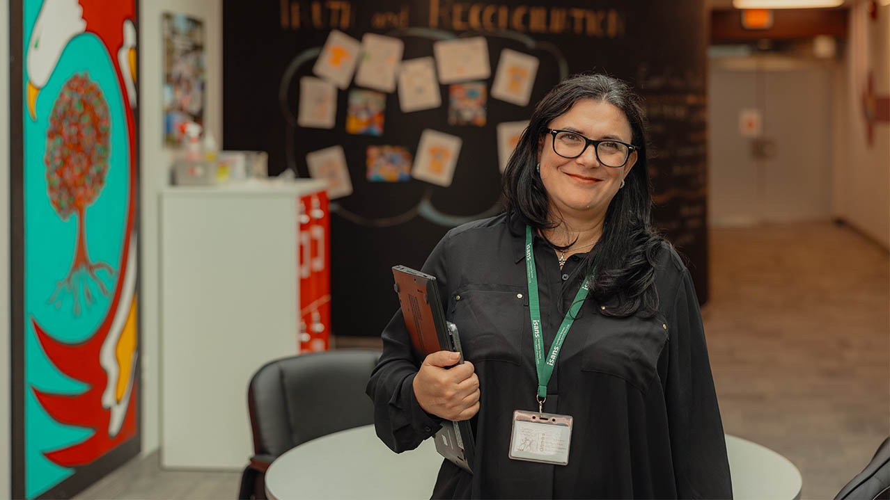 An NSCC Public Relations program graduate stands in front of a table and chairs in an office setting and smiles while holding a laptop.