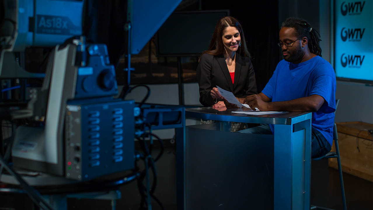 An instructor stands next to a student sitting at a newsdesk in an NSCC studio setting.