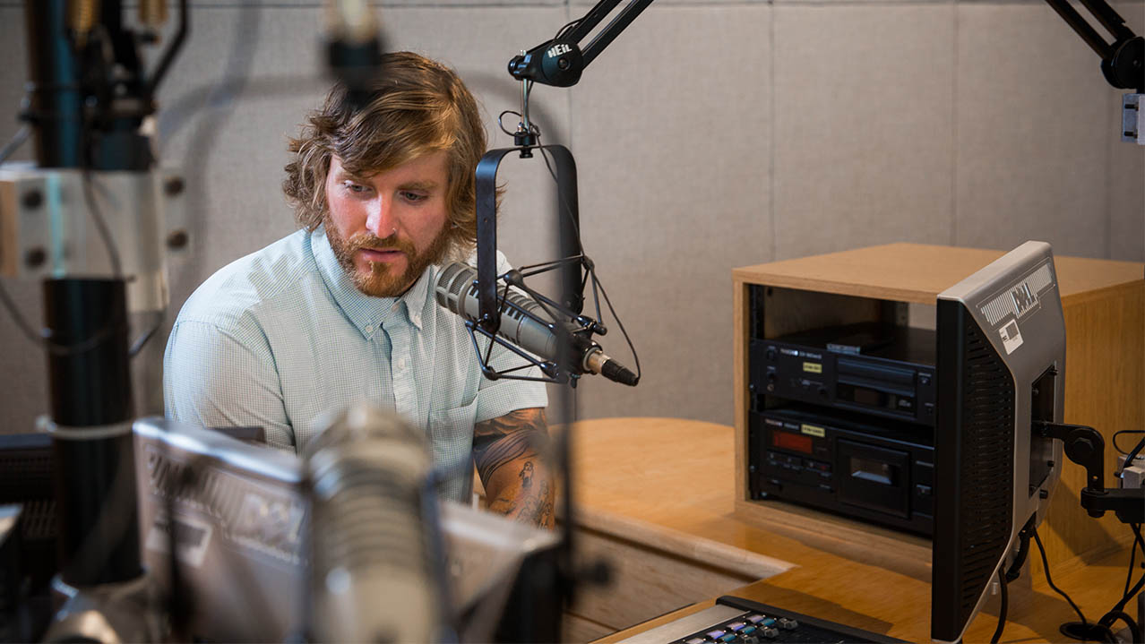 A man sits in front of a mic while seated in a radio studio.