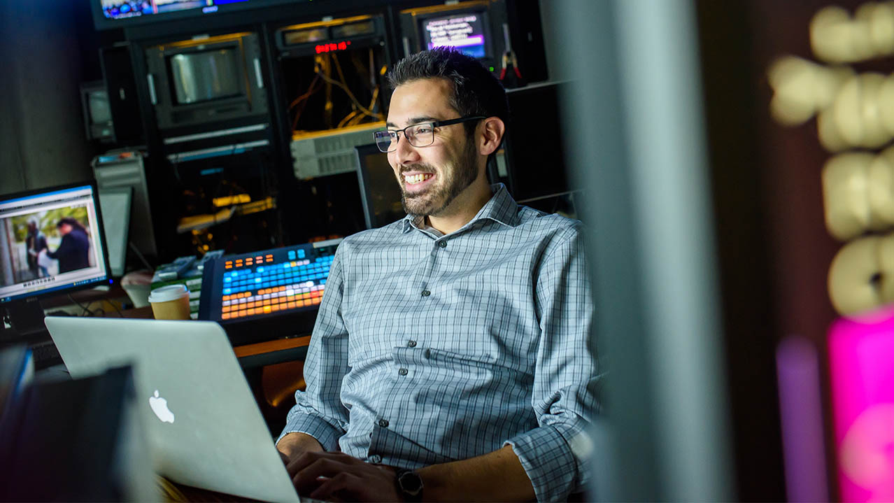 A man sits in a production studio; he's smiling and his laptop in on the desk in front of him.