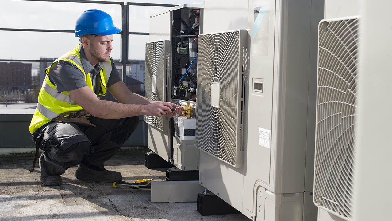 A man in a yellow safety vest and blue hard hat kneels beside an air conditioning unit.