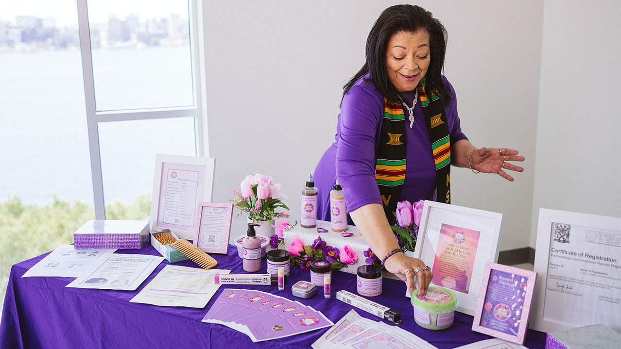 An African Canadian woman smiles as she works on a display of products in decorative jars on a table covered with a purple tablecloth.
