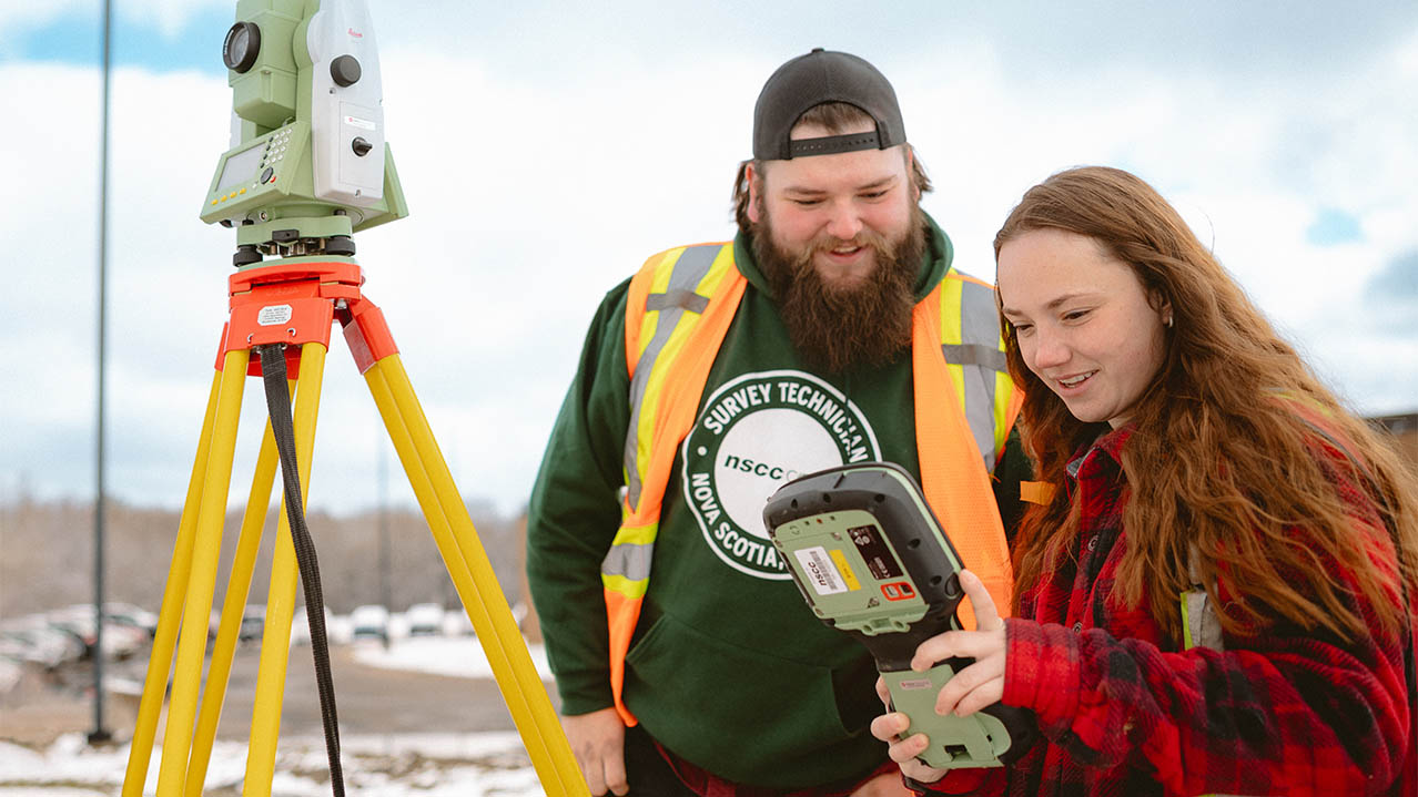 Two students in the Survey Technician program are seen wearing reflective vests, standing outside on a late winter day smiling and looking at a monitor for their surveying equipment.