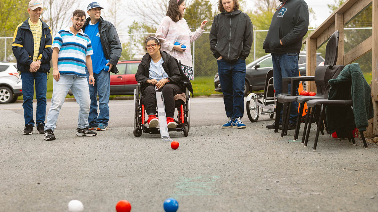 A group of graduates in the Therapeutic Recreation program are seen having fun with their clients outside playing a game with a red ball on the pavement outside a building.