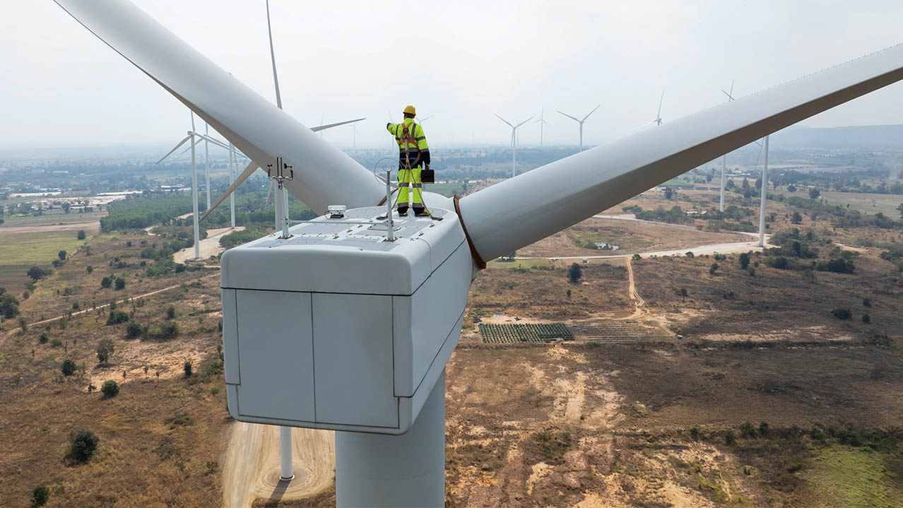 A wind turbine technician stands on top of a wind turbine overlooking a wind farm.