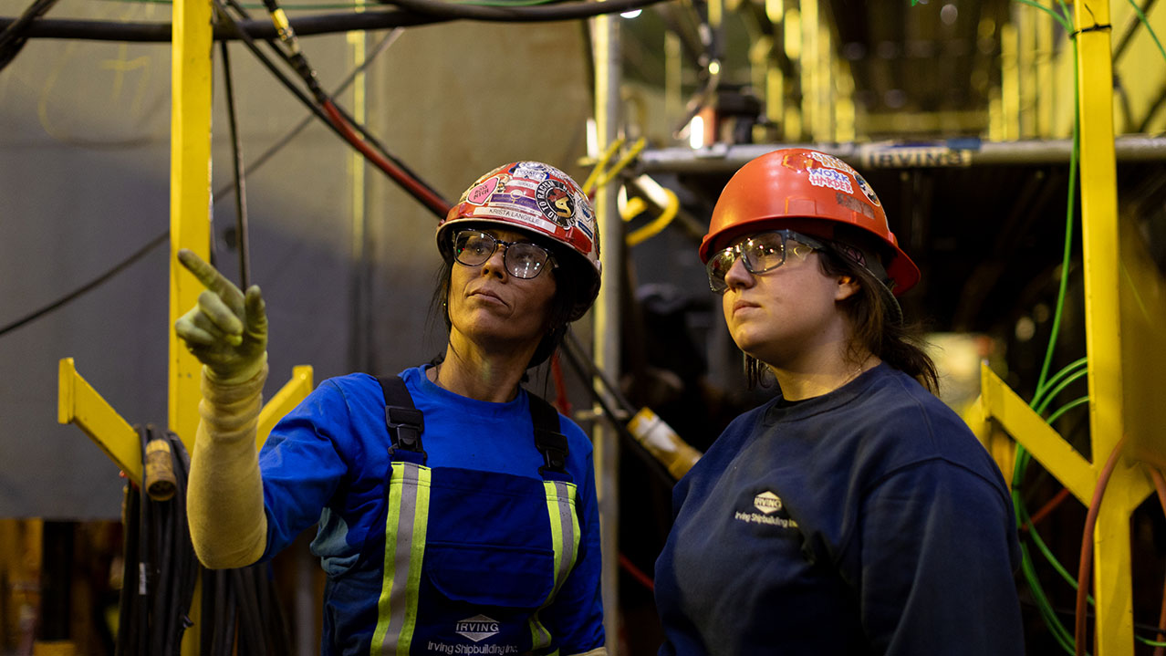 Two NSCC Women Unlimited graduates wearing hard hats and safety glasses stand in an industrial setting.