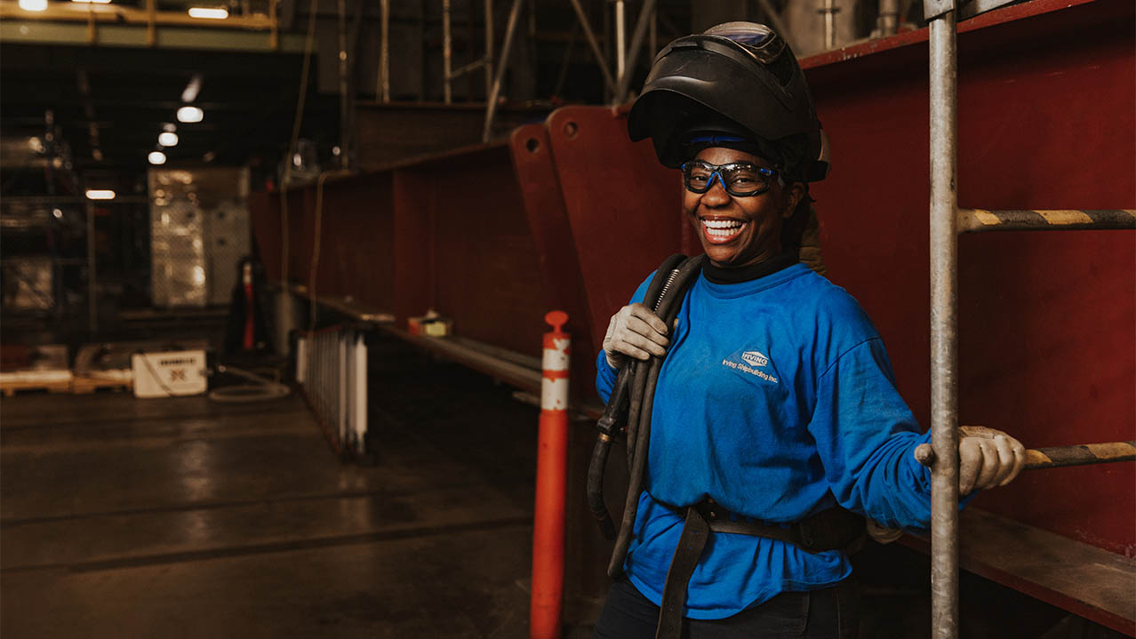 A welder stands in an industrial setting. She wears a long-sleeved shirt, safety goggles, gloves and a welding helmet.