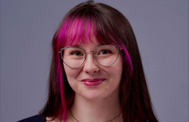 A young woman with round glasses and brown and bright pink hair is seen smiling directly at the camera.