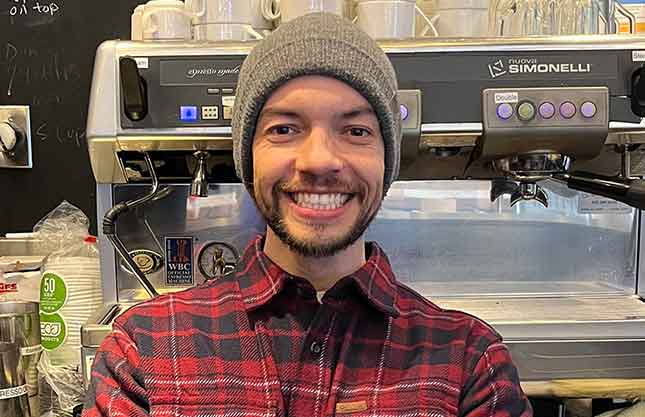 Leo is seen smiling into the camera in front of a cappuccino machine wearing a tan toque.