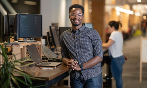 Sandrico, an NSCC graduate, stands in his workplace and leans on a desk while smiling at the camera.