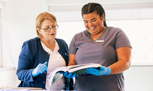 Zena, a Continuing Care grad, stands with an open book and looks at the pages with an instructor to her right.