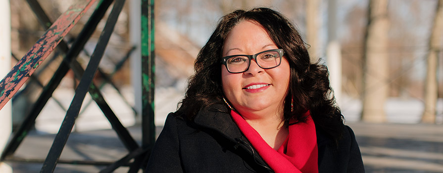 A person with dark, shoulder-length, wavy hair wearing glasses, a coat and a scarf stands outdoors and smiles at the camera.