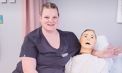 Andrea sits on the edge of a hospital bed next to a mannequin used to practic