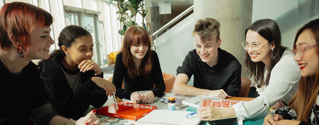 A group of students are seated at a round table, they are looking at a project in the middle of the table.