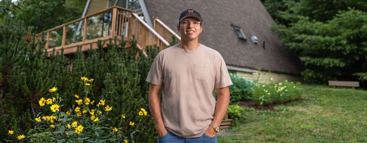 Smiling person standing outdoors in front of a wooden cabin on a sunny day.