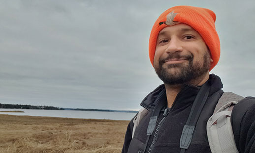 Mitchell stands outdoors near the ocean wearing a hat, jacket and backpack.