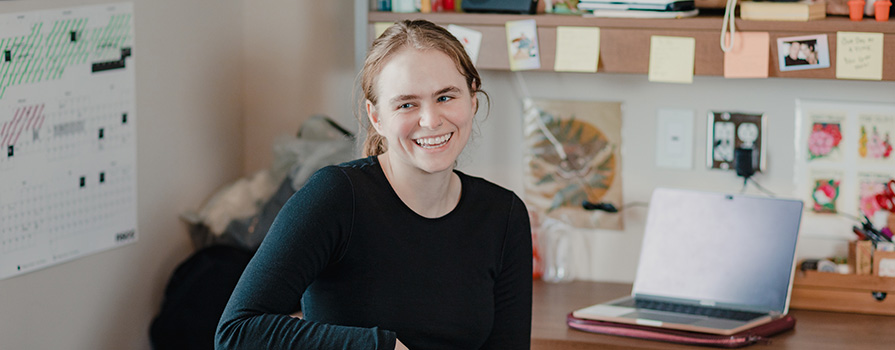 A student smiling, while sitting at a desk with a computer in a background.