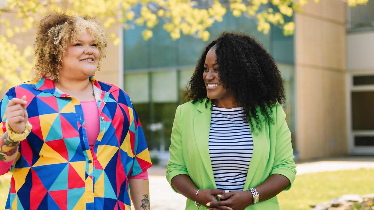 An NSCC student and employee walk side-by-side while talking and smiling outside of an NSCC campus.