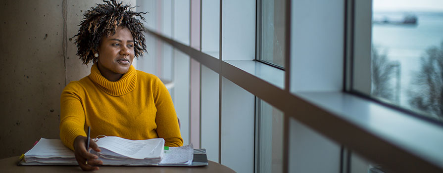 A student sitting at a table with a binder, holding a pen, and glancing out a window.