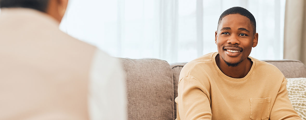 A student sits on a sofa in an office and speaks to someone while smiling.