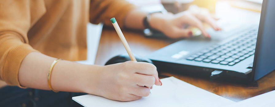 Close-up image of someone taking notes by hand and working on their computer at the same time.