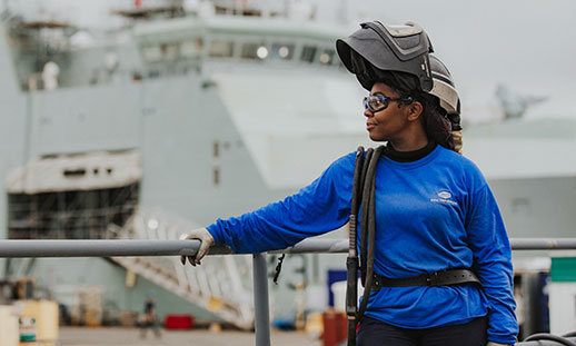 A welder stands in an industrial outdoor setting with ships in the background.