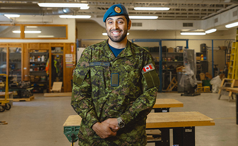 A member of the Canadian Armed Forces stands in uniform in an NSCC workshop and smiles.