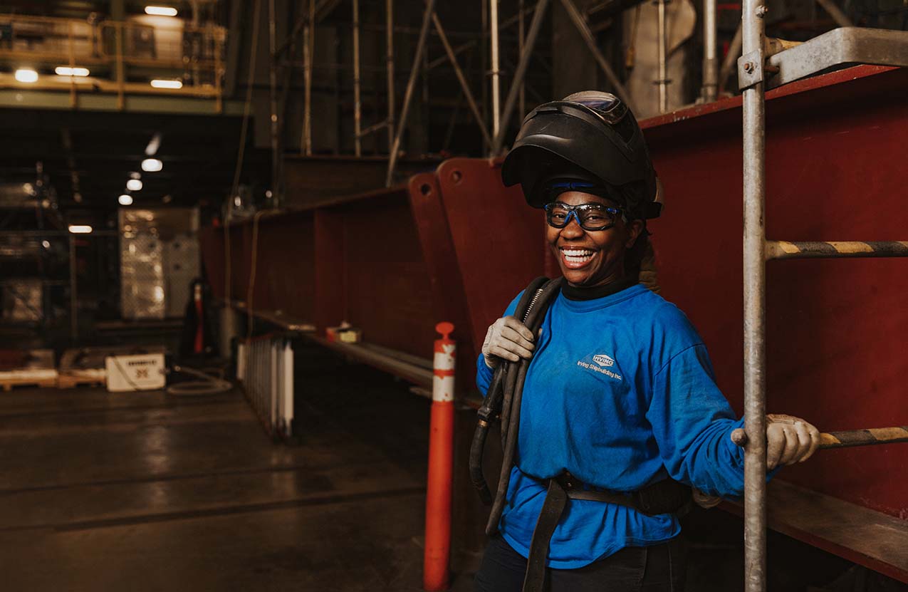 A Black woman in welding gear smiles as she stands in a shop at Halifax Shipyard.