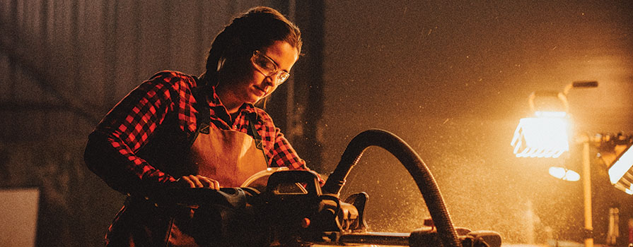 Woman in a plaid shirt wearing safety glasses uses a saw.