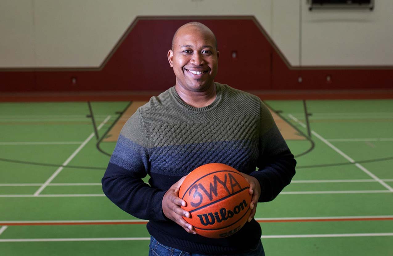 A Black male standing in a gym, smiling, and holding a basketball.