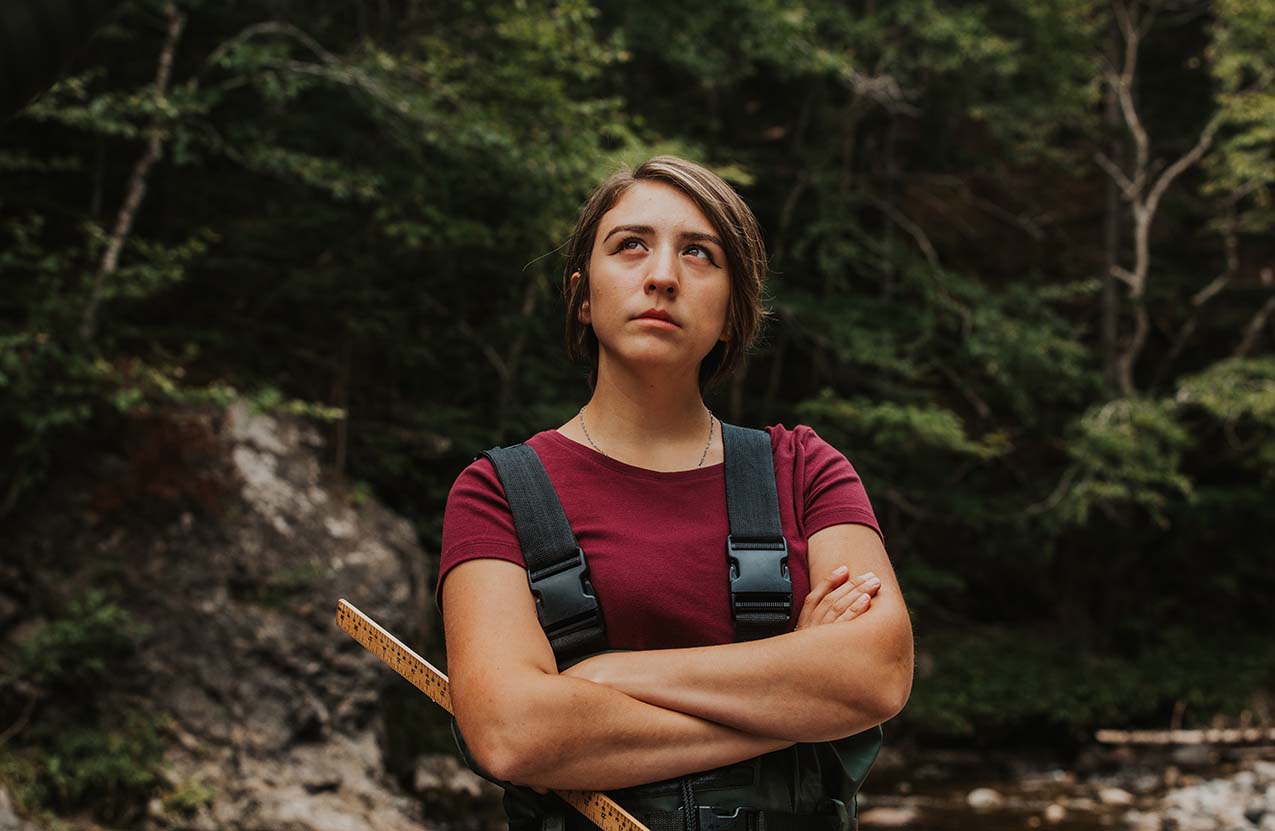 An Indigenous woman, wearing waterproof overalls and arms crossed, looks on.