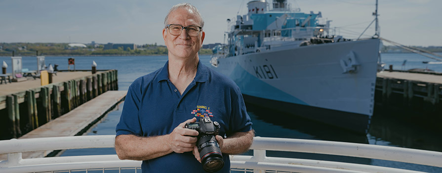 An NSCC graduate and Royal Canadian Navy Veteran stands along the Halifax Waterfront and smiles. He is holding a camera with the Halifax Harbour, two wooden wharves and a ship behind him.