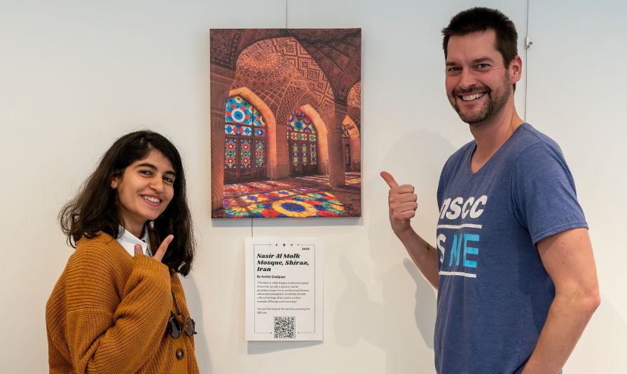 Two people stand and gesture toward an art gallery piece hanging on a wall.
