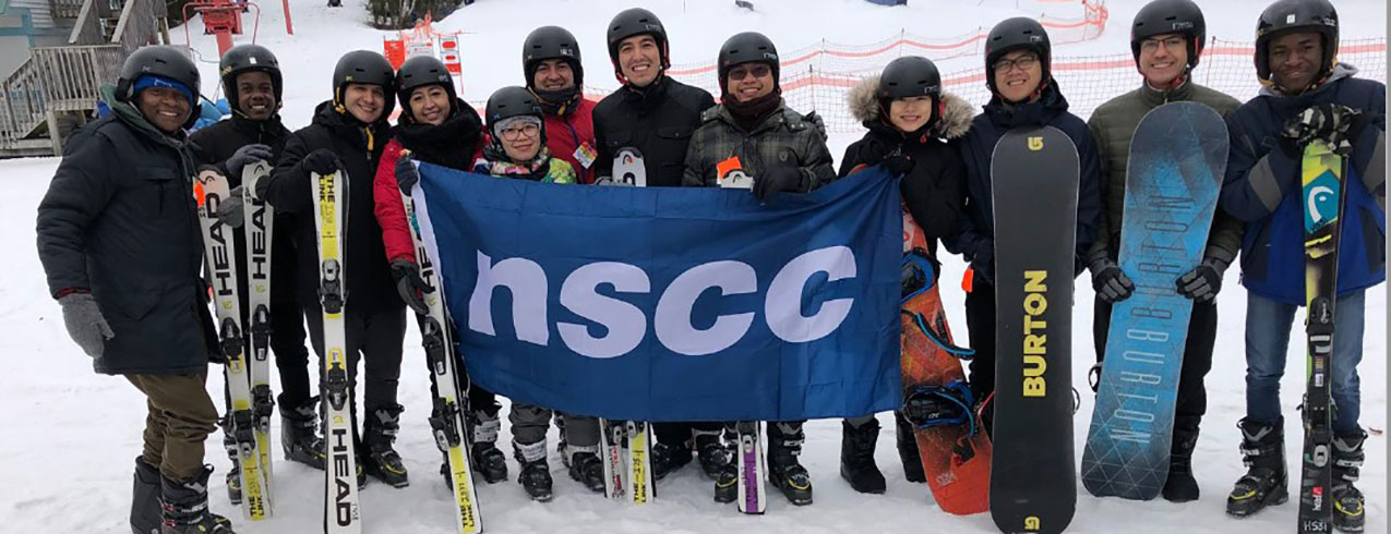 NSCC International students dressed with skiing equipment pose with NSCC flag on snowy slopes in Winter 2019