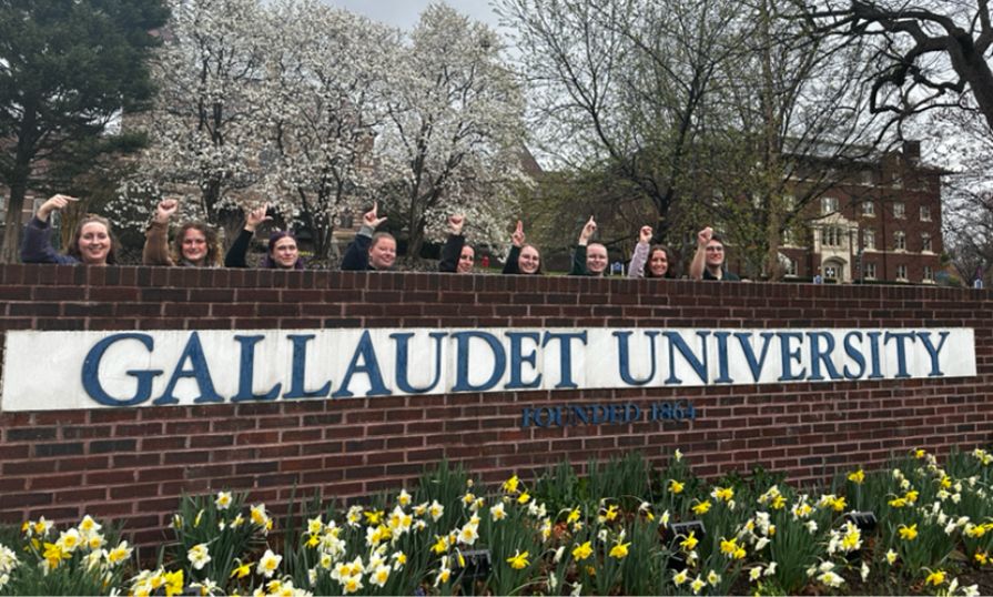 A group of interpreting students poses behind the Gallaudet University sign, raising one finger in celebration, with blooming trees and campus buildings behind them.