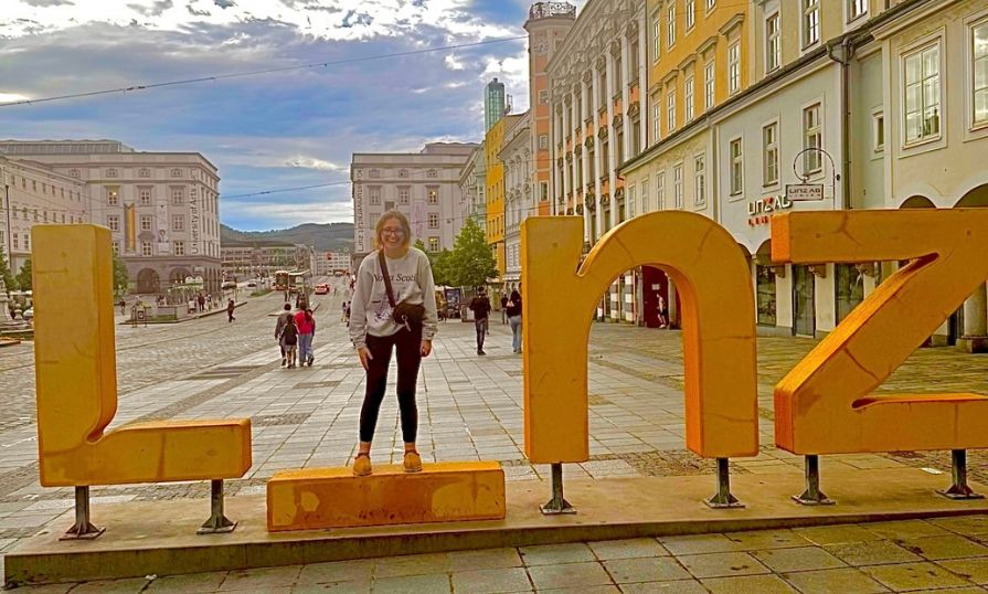 Emily standing inside a large yellow “Linz” sign in a European square, smiling with historic buildings and cloudy sky in the background.