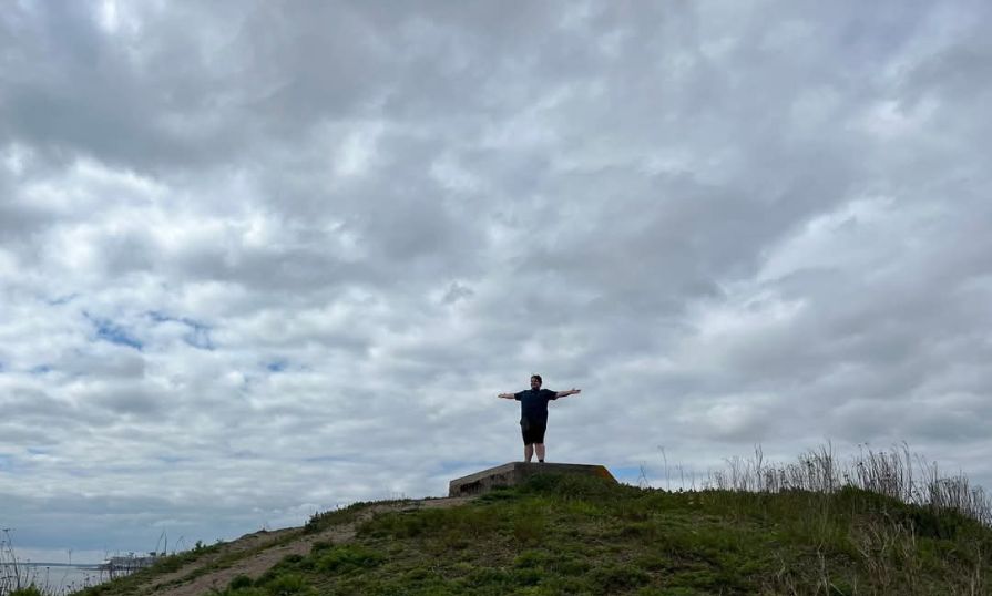 Isaac stands on a grassy hill with his arms stretched out under a cloudy sky