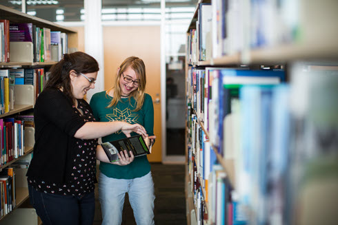 Students standing in library shelves looking at a book. 