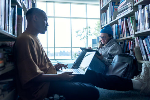 students sitting between library shelves working on laptops