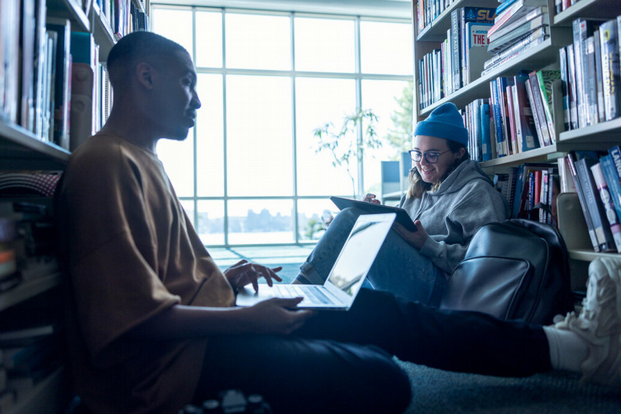 Two students sitting between library stacks working on laptops.